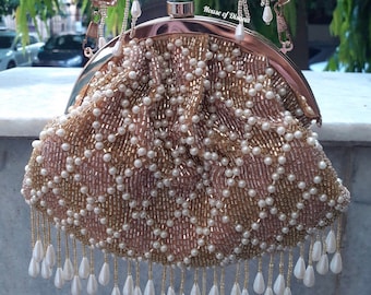May include: A beaded clutch bag with a gold frame and handle. The bag is covered in small, white and gold beads arranged in a diamond pattern. White teardrop-shaped beads hang from the bottom. The text "House of Disana" is visible.
