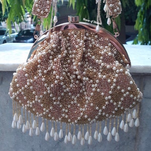 Puede incluir: Un bolso de mano con cuentas, con un marco y asa dorados. El bolso está cubierto de pequeñas cuentas blancas y doradas dispuestas en un patrón de diamantes. Cuentas blancas en forma de lágrima cuelgan de la parte inferior. El texto "House of Disana" es visible.