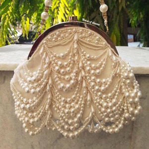 Puede incluir: Un bolso de mano blanco con cierre dorado y una correa de cadena de perlas. El bolso está cubierto de perlas y tiene un diseño en cascada.