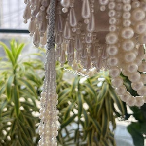 Puede incluir: Primer plano de una l&aacute;mpara de ara&ntilde;a decorativa con cuentas blancas en forma de perlas. Las cuentas est&aacute;n dispuestas en varias formas, incluyendo gotas y hebras. El texto "House of disana" es visible en el lado derecho de la imagen. El fondo presenta plantas verdes y amarillas.