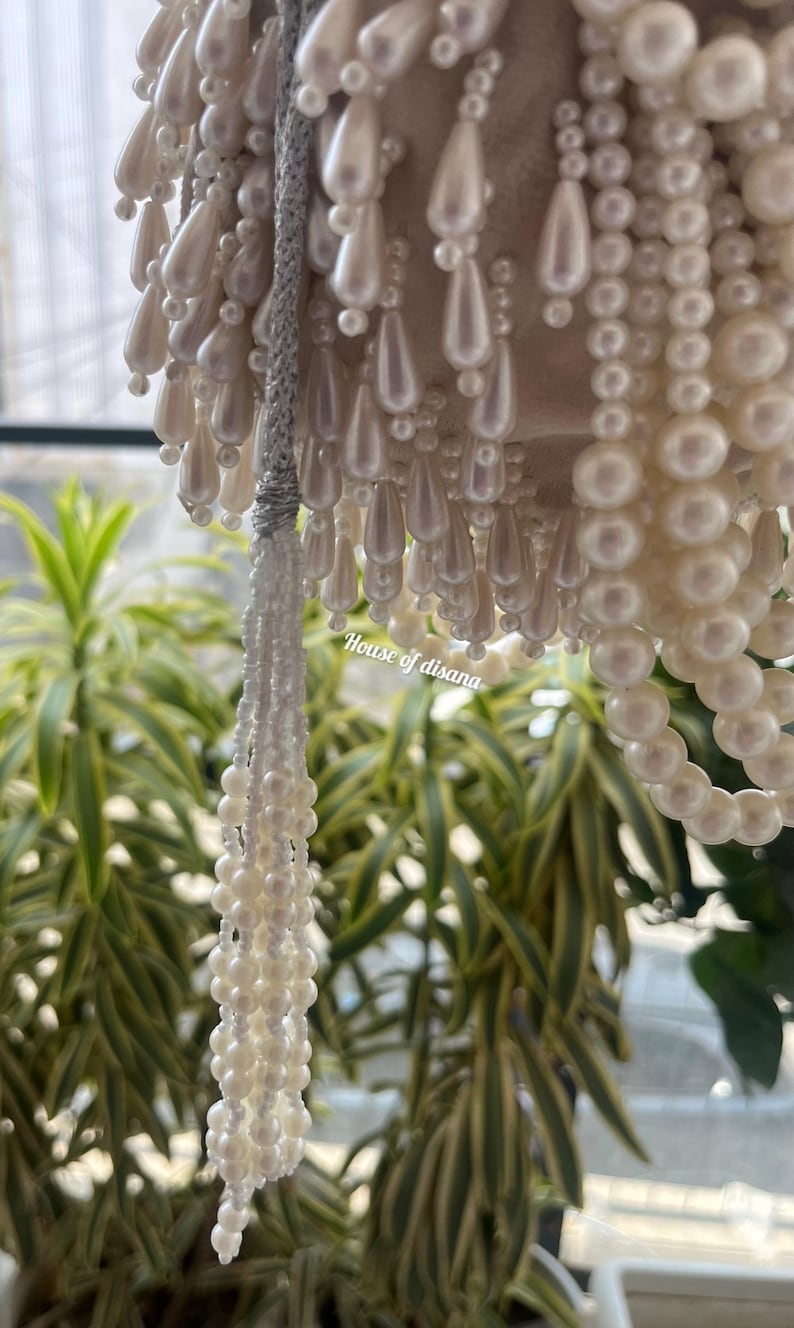 Puede incluir: Primer plano de una l&aacute;mpara de ara&ntilde;a decorativa con cuentas blancas en forma de perlas. Las cuentas est&aacute;n dispuestas en varias formas, incluyendo gotas y hebras. El texto "House of disana" es visible en el lado derecho de la imagen. El fondo presenta plantas verdes y amarillas.