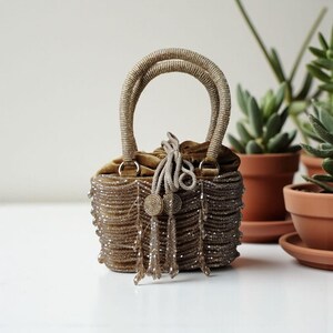 Op de afbeelding: Een kleine, met kralen bezette handtas met een fluwelen trekkoordsluiting en met kralen bezette handvatten. De tas is lichtbruin van kleur met bungelende kralenfranjes en decoratieve kwastjes. De tas staat op een wit oppervlak met potplanten op de achtergrond.