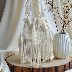 Può includere: Borsa a coulisse con perline color avorio con lunghe frange. La borsa ha un manico in raso bianco ed è decorata con piccole perle. La borsa è esposta su una fetta di legno con un vaso bianco e una composizione floreale sullo sfondo.