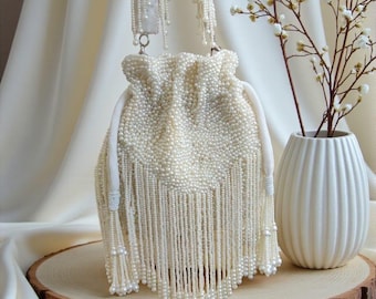 May include: Ivory beaded drawstring bag with long fringe. The bag has a white satin handle and is decorated with small pearls. The bag is displayed on a wooden slice with a white vase and floral arrangement in the background.
