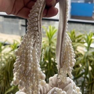 Puede incluir: Bolso de mano color crema adornado con numerosas perlas de imitaci&oacute;n. El asa y el cuerpo del bolso est&aacute;n adornados con perlas de varios tama&ntilde;os, creando una apariencia lujosa y texturizada. El dise&ntilde;o del bolso es elegante y adecuado para ocasiones especiales.