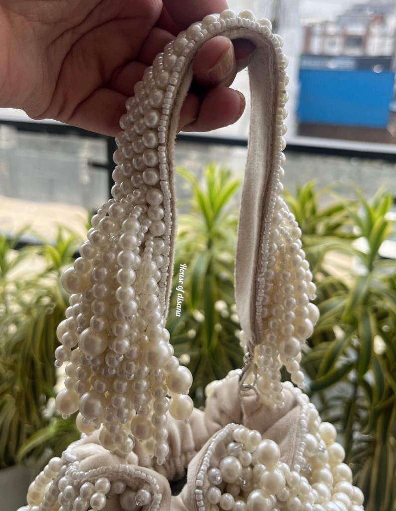 Puede incluir: Bolso de mano color crema adornado con numerosas perlas de imitaci&oacute;n. El asa y el cuerpo del bolso est&aacute;n adornados con perlas de varios tama&ntilde;os, creando una apariencia lujosa y texturizada. El dise&ntilde;o del bolso es elegante y adecuado para ocasiones especiales.