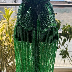 Pode incluir: Uma bolsa verde com contas e cordão, com longas franjas. A bolsa é adornada com pequenas contas verdes brilhantes e tem um fecho de cordão verde escuro. As franjas pendem da parte inferior, adicionando um elemento decorativo.