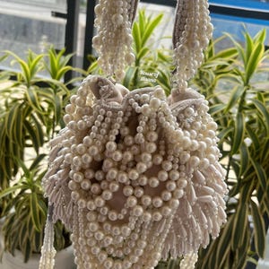 Puede incluir: Bolso de mano con cuentas de color marfil con un interior suave de color rubor. La bolsa est&aacute; muy adornada con perlas de imitaci&oacute;n de varios tama&ntilde;os, creando una apariencia lujosa y texturizada. Las asas y las borlas tambi&eacute;n est&aacute;n adornadas con cuentas. El texto "House of disana" es visible.