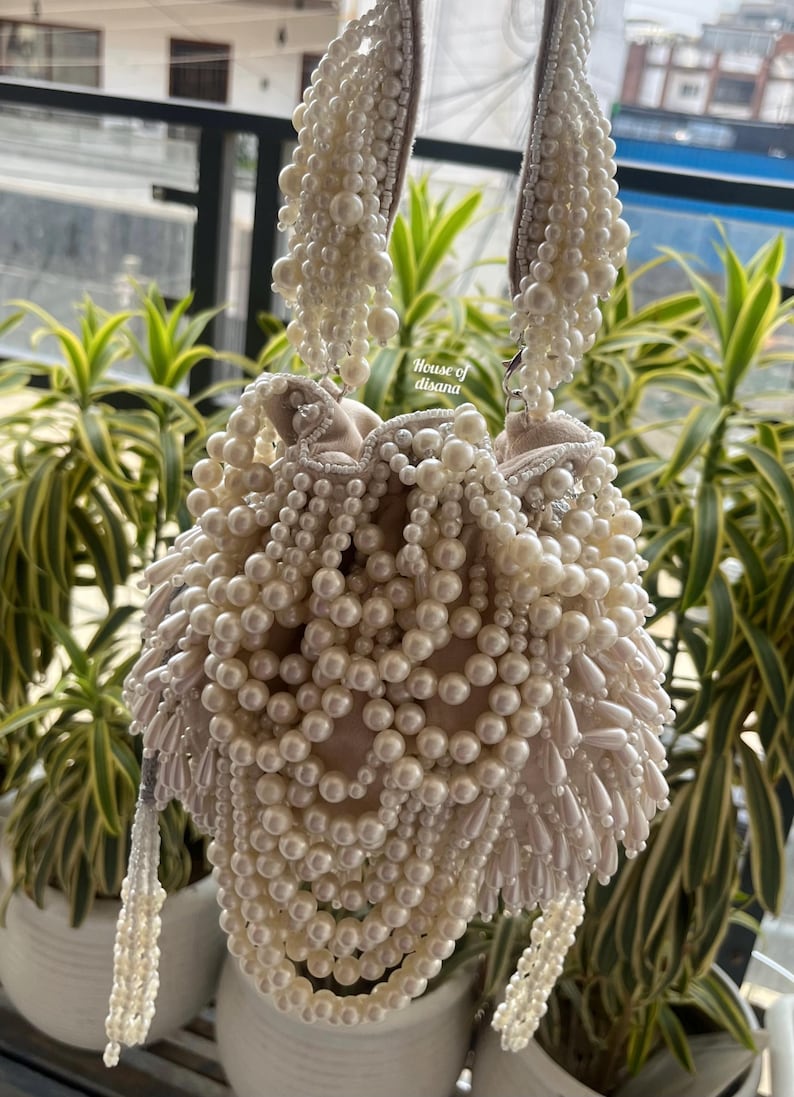 Puede incluir: Bolso de mano con cuentas de color marfil con un interior suave de color rubor. La bolsa est&aacute; muy adornada con perlas de imitaci&oacute;n de varios tama&ntilde;os, creando una apariencia lujosa y texturizada. Las asas y las borlas tambi&eacute;n est&aacute;n adornadas con cuentas. El texto "House of disana" es visible.