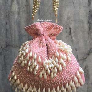 Könnte beinhalten: Eine rosa Samt-Kordeltasche mit einem goldenen Kettengriff und einem Fransenbesatz aus weißen Perlen. Die Tasche ist mit weißen Perlen verziert.