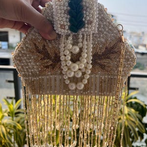 May include: An ornate, cream-colored beaded clutch with gold embroidery and a dangling fringe. The clutch features a decorative green accent and a gold chain strap. The design includes pearl and bead embellishments, creating a luxurious aesthetic.