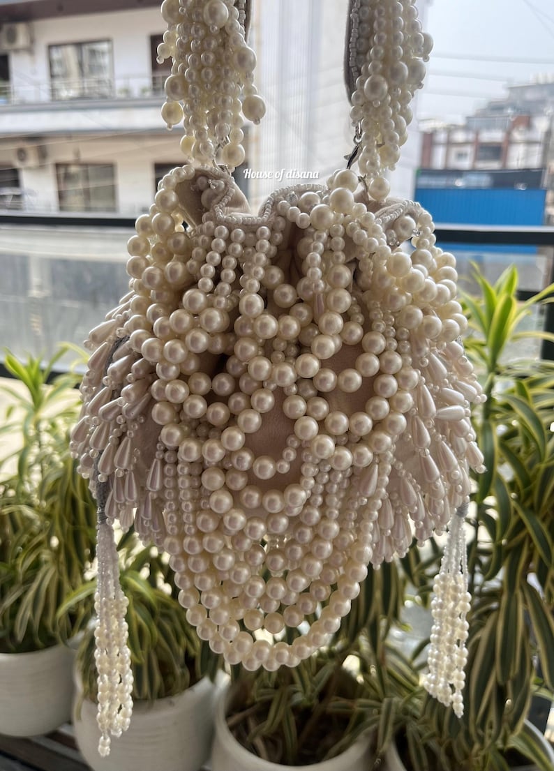 Puede incluir: Un bolso de mano con cuentas de color crema con cierre de cord&oacute;n. El bolso est&aacute; muy adornado con perlas de imitaci&oacute;n de varios tama&ntilde;os, creando una apariencia texturizada y elegante. El texto "House of aisana" es visible.