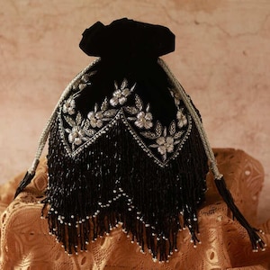May include: Black beaded evening bag with silver floral embroidery. The bag has a drawstring closure and long beaded fringe. The straps are silver and black. The bag is on a patterned surface.