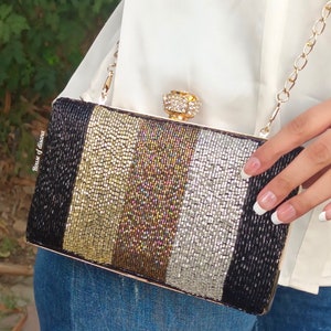 Puede incluir: Un bolso de mano negro, dorado y plateado con cuentas, con un cierre dorado y un adorno de cristal. El bolso de mano tiene una correa de cadena dorada.