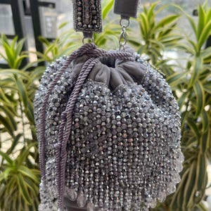 May include: A gray beaded drawstring bag with a matching strap. The bag is covered in small, shiny beads, with a beaded fringe at the bottom. The strap and drawstring are a lighter shade of gray. The bag is hanging in front of green plants.
