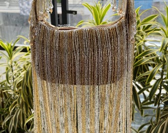 May include: A beaded handbag with a curved top and a long fringe. The bag is primarily gold, with accents of silver and brown. The handle is a matching gold color, and the fringe hangs down from the bottom of the bag.