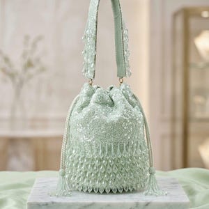 Puede incluir: Un bolso tipo saco de color verde menta claro con cierre de cordón, cubierto totalmente de lentejuelas brillantes. El bolso está adornado con cuentas translúcidas en forma de lágrima en la base y el asa, con borlas de cuentas.