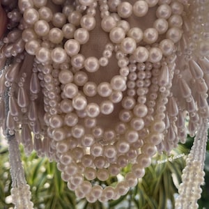 Puede incluir: Bolso de mano con cuentas de color marfil con un interior suave y claro. El bolso est&aacute; adornado con m&uacute;ltiples hilos de perlas redondas y brillantes y cuentas en forma de l&aacute;grima, creando una superficie lujosa y texturizada. El bolso tiene un asa con cuentas.