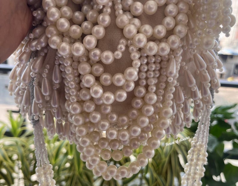 Puede incluir: Bolso de mano con cuentas de color marfil con un interior suave y claro. El bolso est&aacute; adornado con m&uacute;ltiples hilos de perlas redondas y brillantes y cuentas en forma de l&aacute;grima, creando una superficie lujosa y texturizada. El bolso tiene un asa con cuentas.