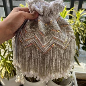May include: A silver beaded drawstring pouch with a chevron pattern. The pouch features a white fabric top and is adorned with silver beads, sequins, and dangling pearl-like accents. The bag is designed for carrying small items.