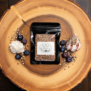 May include: A black resealable pouch with a white label reading Sprout and Shore, containing a textured brown mixture. The pouch is surrounded by fresh blueberries, a white spiral seashell, and a patterned conical seashell on a wood slice.