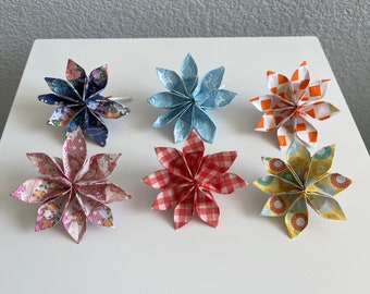 May include: Six origami flowers in various colors and patterns. The flowers are made of paper and have a starburst shape. The flowers are arranged in a row on a white surface.