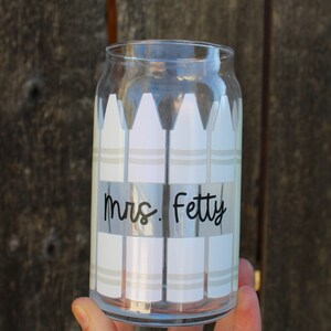 16 oz clear glass can with lined up vertically  crayons that are white since they're room temperature, wrapped around the cup, rectangle cutout in the middle of 4 front crayons with black cursive teacher name