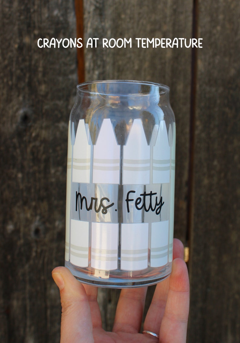 16 oz clear glass can with lined up vertically  crayons that are white since they're room temperature, wrapped around the cup, rectangle cutout in the middle of 4 front crayons with black cursive teacher name