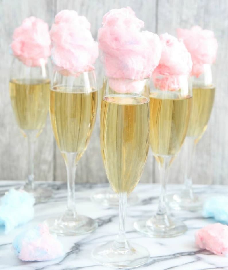 May include: Six champagne flutes filled with a pale yellow liquid, each topped with a pink cotton candy cloud. The glasses are on a white marble surface.