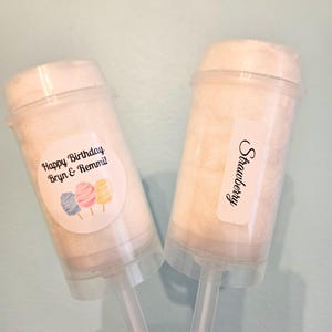 May include: Two clear plastic push-pop containers filled with white cotton candy. One container has a sticker that says "Happy Birthday Bryn & Remmi!" with three cartoon cotton candy cones. The other container has a sticker that says "Strawberry".