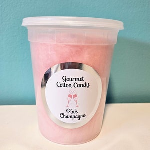 May include: A clear plastic container filled with pink cotton candy. The container has a silver label that reads "Gourmet Cotton Candy" and "Pink Champagne".
