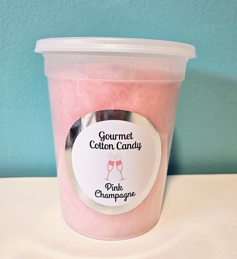 May include: A clear plastic container filled with pink cotton candy. The container has a silver label that reads "Gourmet Cotton Candy" and "Pink Champagne".