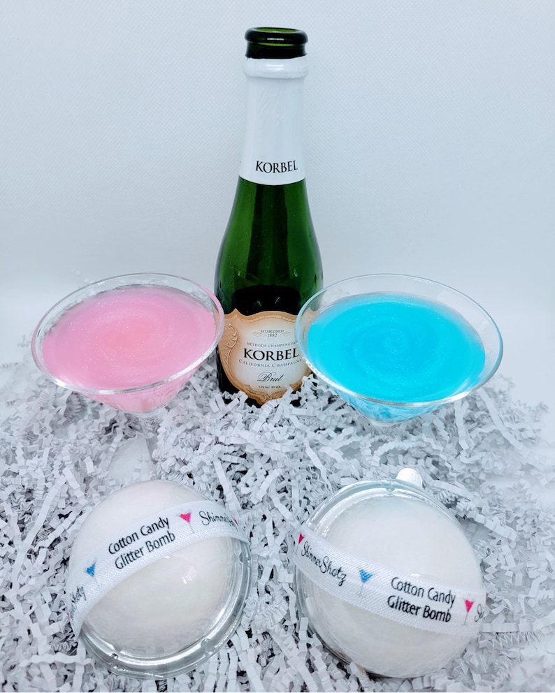 K&ouml;nnte beinhalten: Zwei wei&szlig;e Badebomben mit einem blauen und rosa Band, auf dem "Cotton Candy Glitter Bomb" und "Shimmer Shots" steht, sowie eine Champagnerflasche und zwei Martini-Gl&auml;ser, gef&uuml;llt mit rosa und blauer Fl&uuml;ssigkeit.