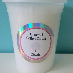 May include: A white plastic container with a lid, filled with white cotton candy. The container has a holographic sticker with the text "Gourmet Cotton Candy" and "Classic" in pink and white.