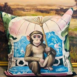 May include: Cushion with a print of a monkey holding a parasol. The monkey is brown and seated on a blue bench with floral patterns. The parasol is pink and white, and the cushion has green leaf accents.
