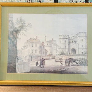 May include: Framed watercolor print depicting a European town scene. The artwork features buildings, people, and horses pulling a cart on a cobblestone street. The print is set in a gold frame with a green mat.