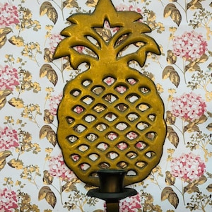 May include: A gold-colored pineapple-shaped wall sconce with a candle holder. The sconce is made of metal and has a detailed design. The background is a floral wallpaper with pink and gold accents.