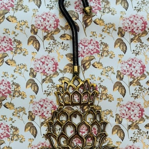 May include: A decorative brass pineapple-shaped object with a black handle. The pineapple has a detailed, openwork design. The background is a floral pattern with pink and gold flowers on a white background.