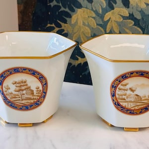 May include: Two white hexagonal porcelain planters featuring a gold rim and base. Each planter displays a central circular medallion with a sepia-toned landscape illustration of a building and trees, framed by a decorative blue and gold border.