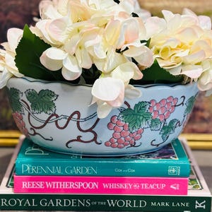 May include: A floral arrangement featuring white and pink hydrangeas in a decorative bowl with a grape design, resting on a stack of books. The books include "Perennial Garden", "Reese Witherspoon Whiskey Teacup", and "Royal Gardens of the World".