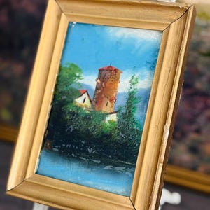 May include: Small framed painting with a gold-colored wooden frame. The artwork depicts a tower with a red roof, surrounded by green trees and a blue sky. The painting is displayed on a white decorative stand.