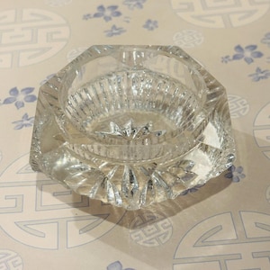 May include: A clear, faceted glass ashtray with a circular inner bowl and a starburst design on the base. The ashtray has an octagonal shape and is set against a patterned background.