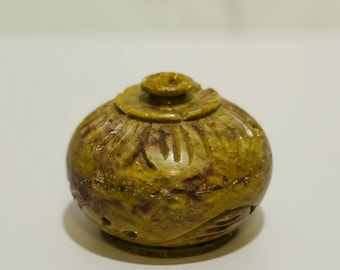 May include: A small, round, olive-green stone container with a lid. The container has carved floral patterns and a small knob on top. The stone has variations in color, with darker brown and lighter yellow-green hues.