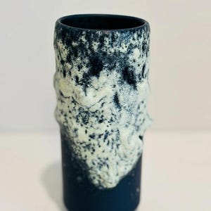 May include: A dark blue ceramic vase with a white textured design. The vase has a cylindrical shape and a smooth, rounded rim.