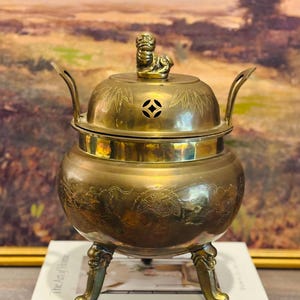 May include: A vintage brass incense burner with a lid, decorative handles, and three ornate feet. The lid is topped with a small figurine. The burner is sitting on top of two books, "The Art of Home" and "Living with BLUE & WHITE".