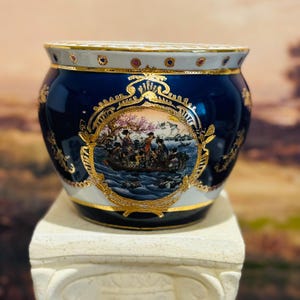 May include: A decorative ceramic planter with a navy blue base, white rim, and gold accents. The planter features a detailed illustration of a boat scene within a gold frame. The planter is displayed on a white pedestal.