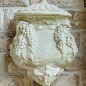 May include: A white ceramic wall-mounted container with a lid and grapevine designs. The container has an iridescent finish.