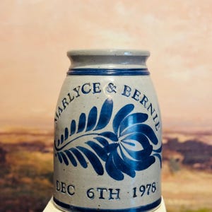 May include: A vintage stoneware jar with a gray and blue color scheme. The jar features the names "Marlyce & Bernie" and the date "DEC 6TH 1978" in blue lettering, along with a blue floral design. The jar has a cylindrical shape.