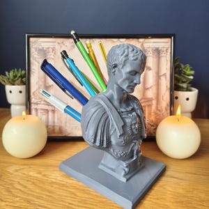 May include: A grey pen holder in the form of a Roman emperor bust, filled with pens and pencils of various colours. The holder sits on a square base. The background features a framed picture, candles, and small potted plants on a wooden table.