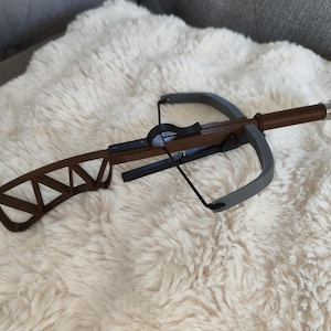 May include: A miniature crossbow with a brown wooden body, gray limbs, and a black string. The handle is wrapped in brown material, and the bow is resting on a fluffy, white surface.